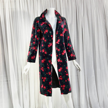 May include: A long-sleeved black coat featuring a vibrant all-over print of red roses with green leaves. The coat has a structured collar, a button-front closure, and a knee-length hemline, displayed on a white mannequin against a white curtain.